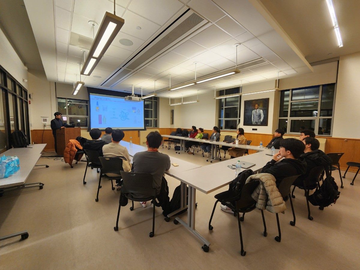 A speaker at a lectern presenting polymer chemistry slides to attendees seated around a long table in Olin Hall