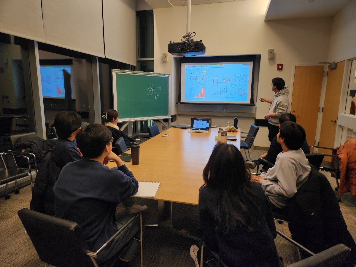 A speaker beside a projected chemistry slide and a chalkboard showing a molecular structure, with attendees around a seminar table