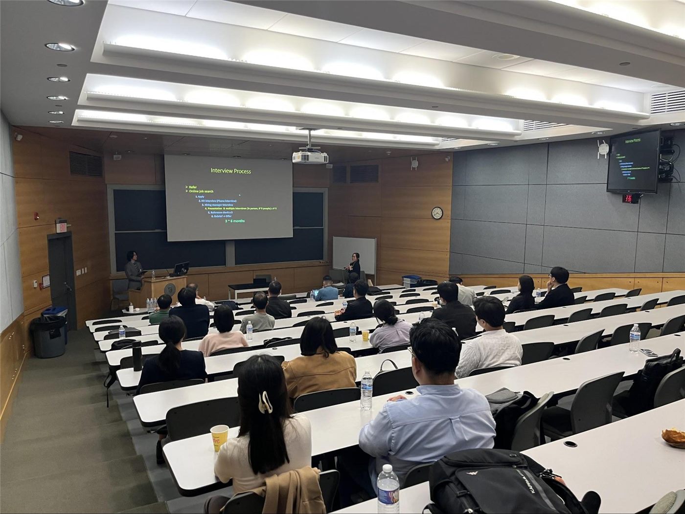 A speaker presenting a slide titled Interview Process to a seated audience in a tiered lecture hall