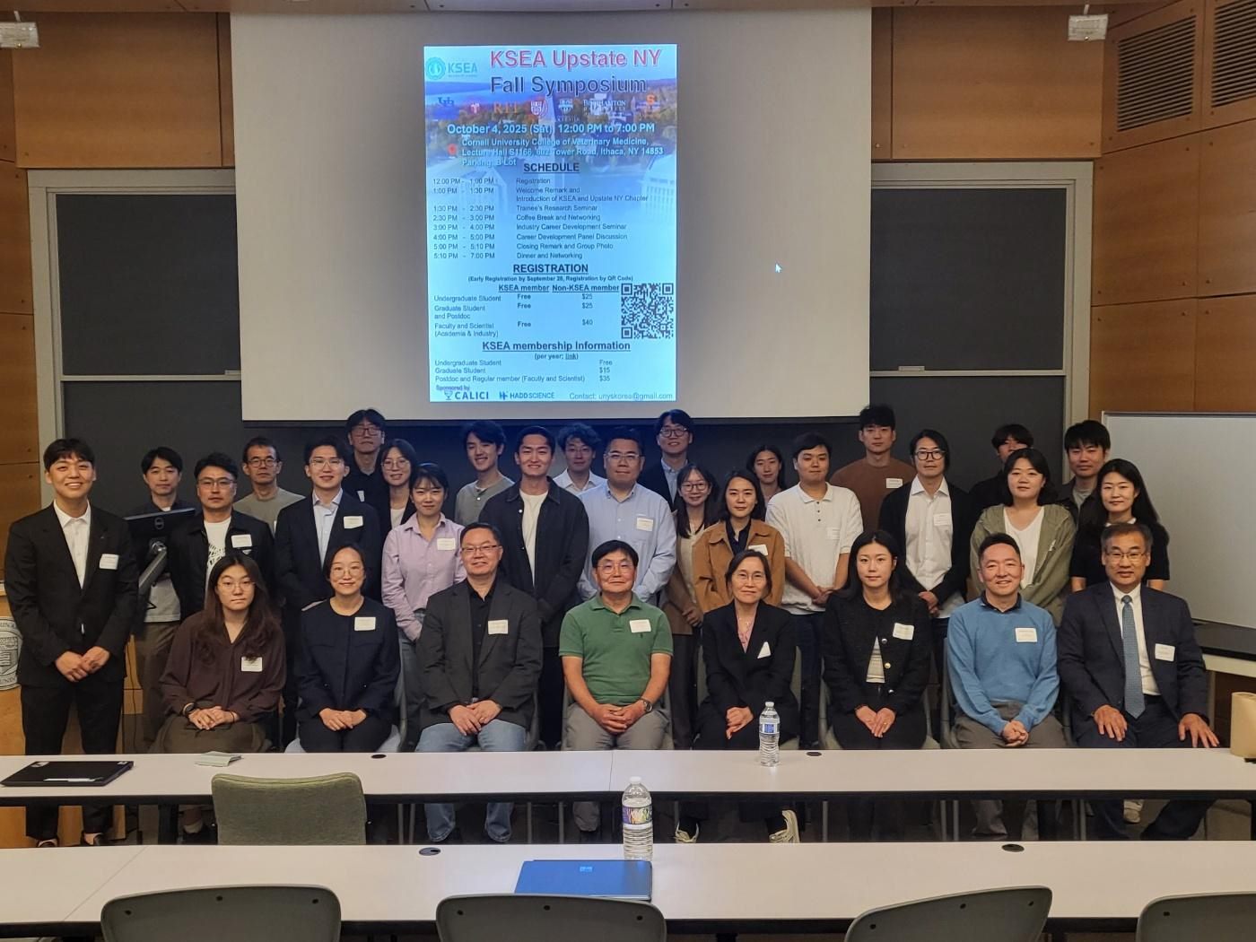 Roughly forty attendees posed together in a Cornell lecture hall beneath the Fall Symposium programme slide