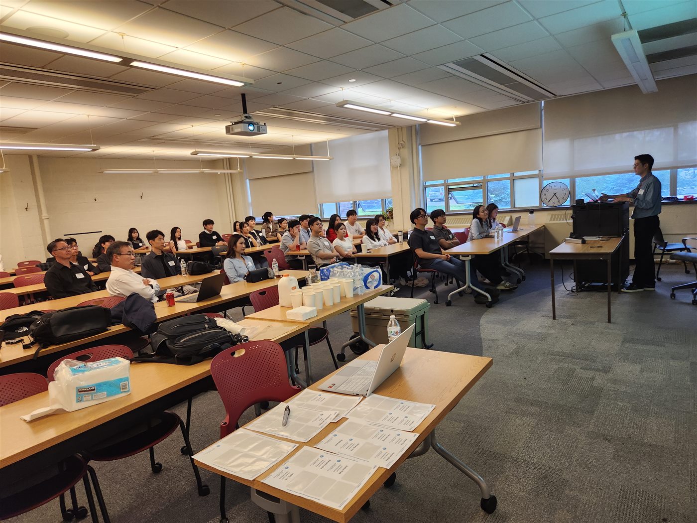 A speaker presenting to attendees seated at tables in a classroom, with printed handouts and refreshments on the tables